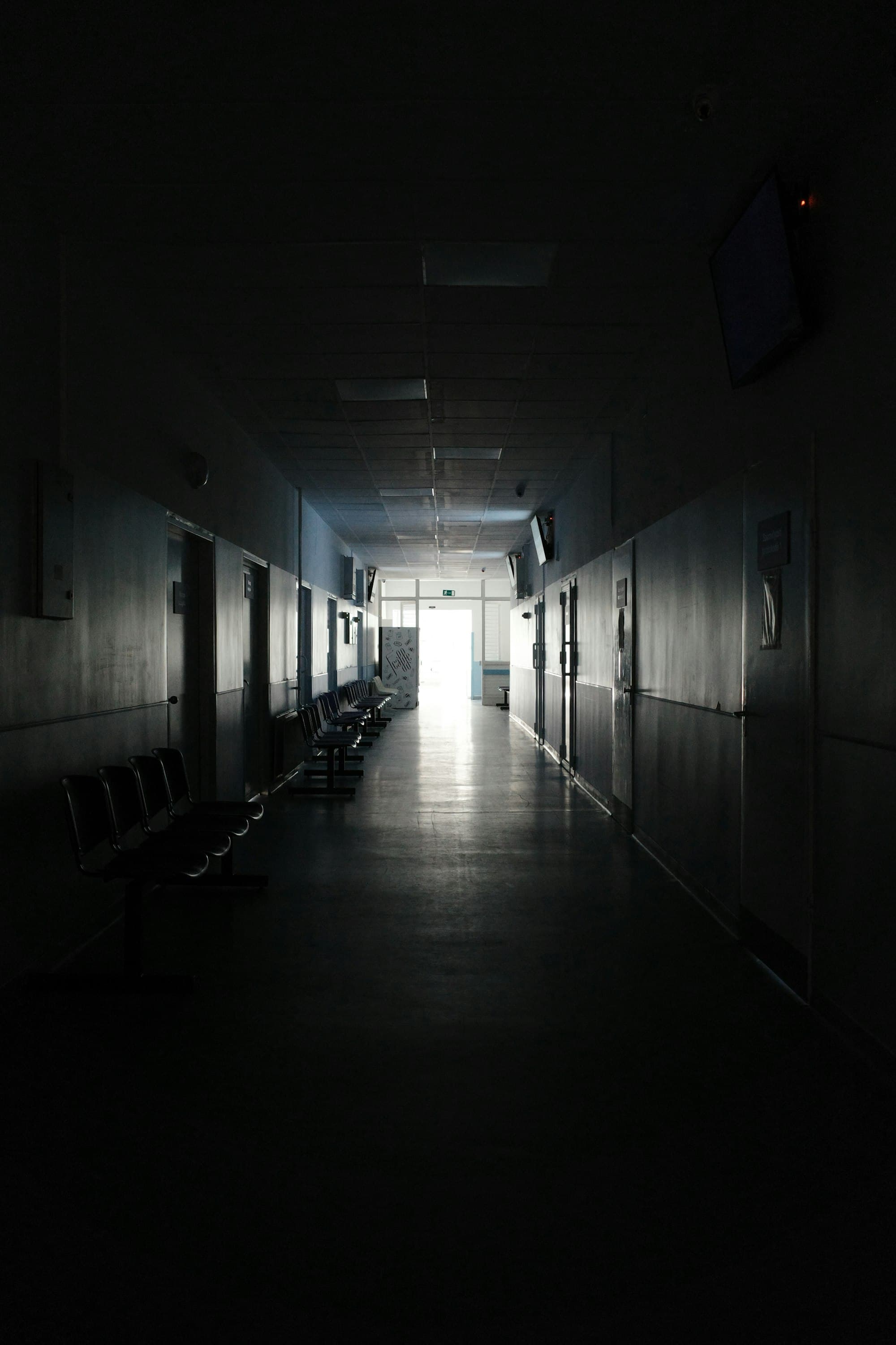 A dark hospital corridor with benches, leading toward a lit doorway.