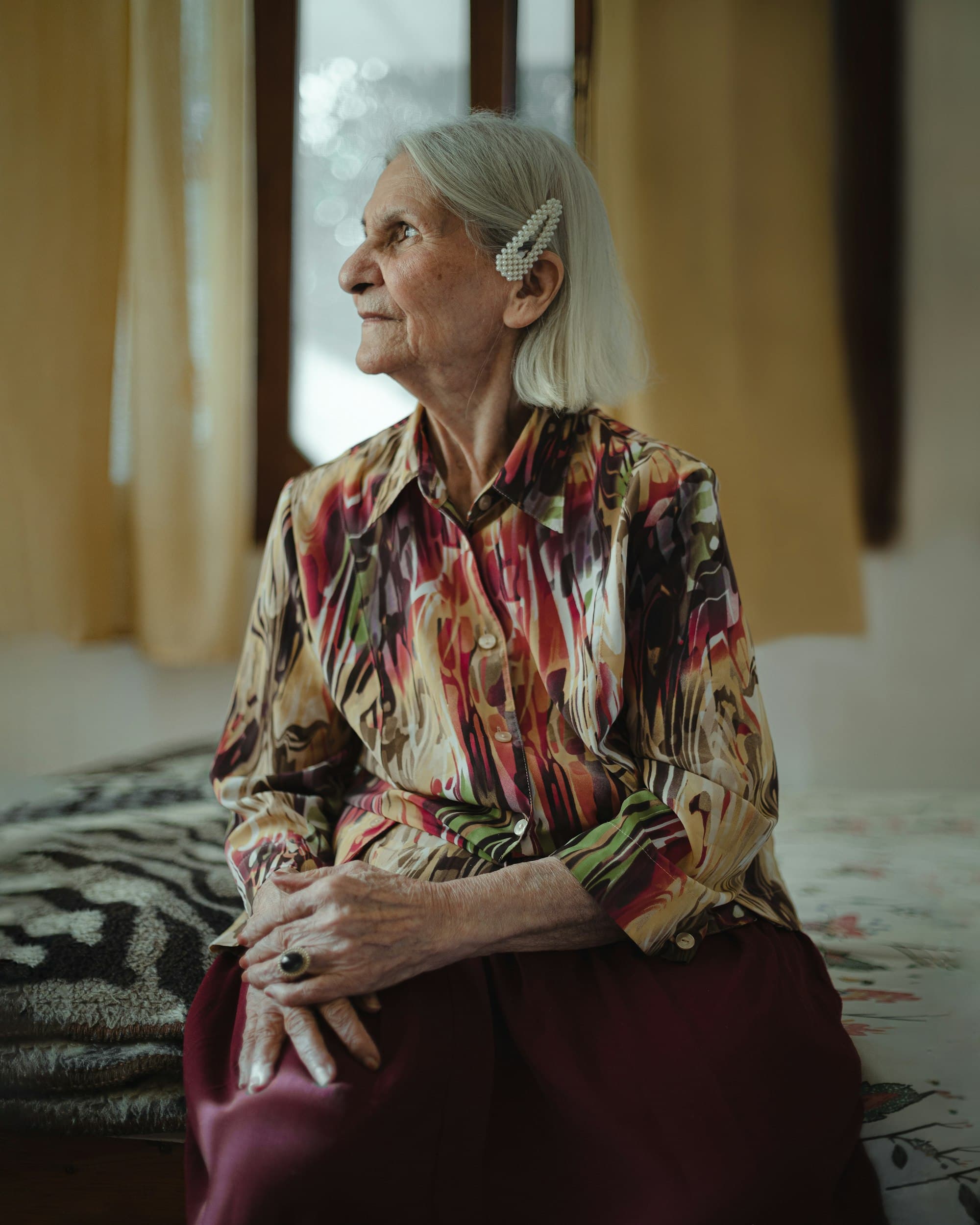 An older woman sitting quietly on her bed at home, hands folded.